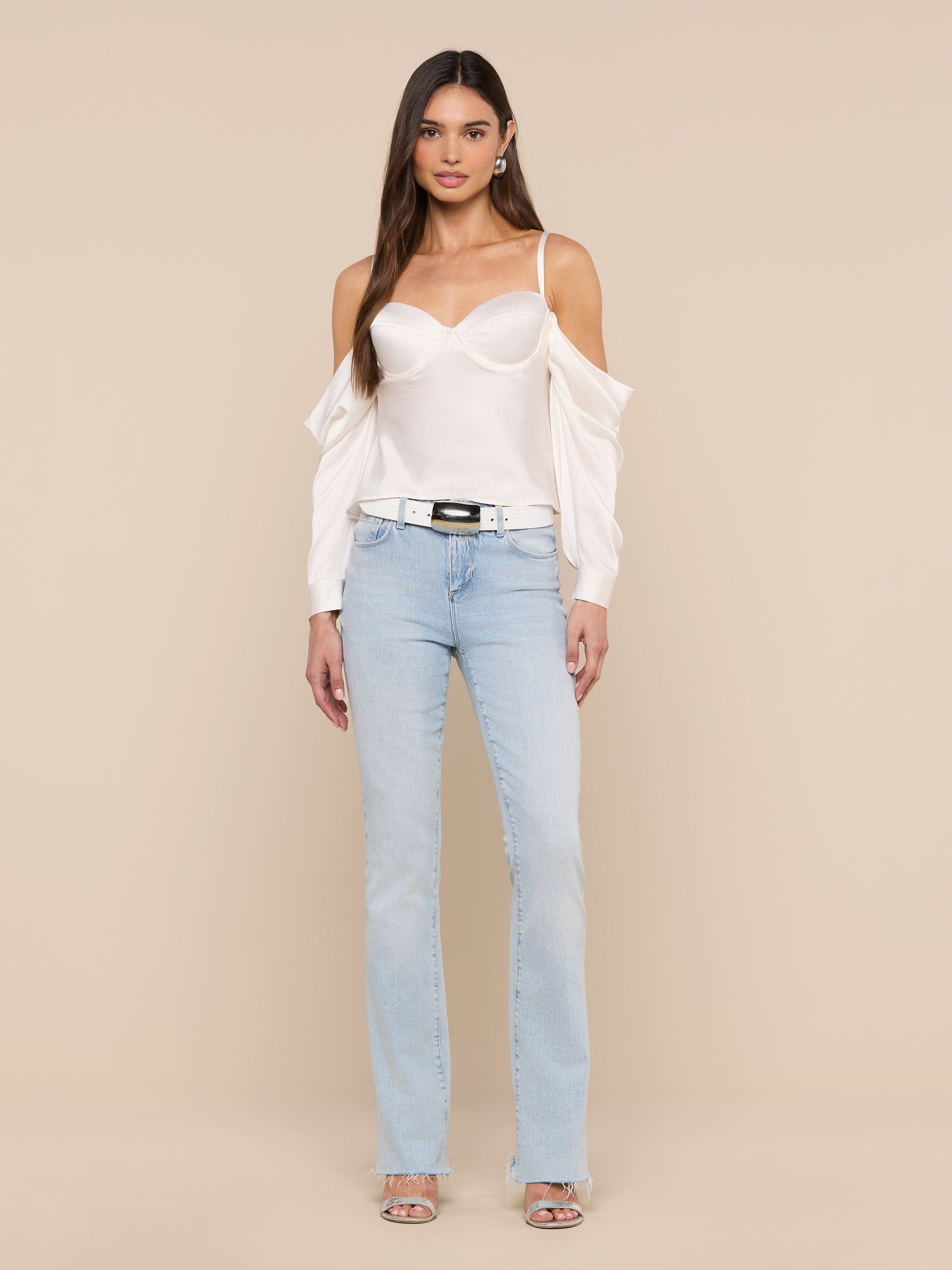 A woman stands against a plain white background wearing the Saya Satin Top in white, light blue jeans, a black belt, and open-toe heels. Her arms are relaxed by her sides and her long, straight hair is loose.