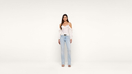 A woman stands against a plain white background, wearing a white off-shoulder blouse, light blue jeans, and heeled sandals. She looks directly at the camera with a neutral expression.