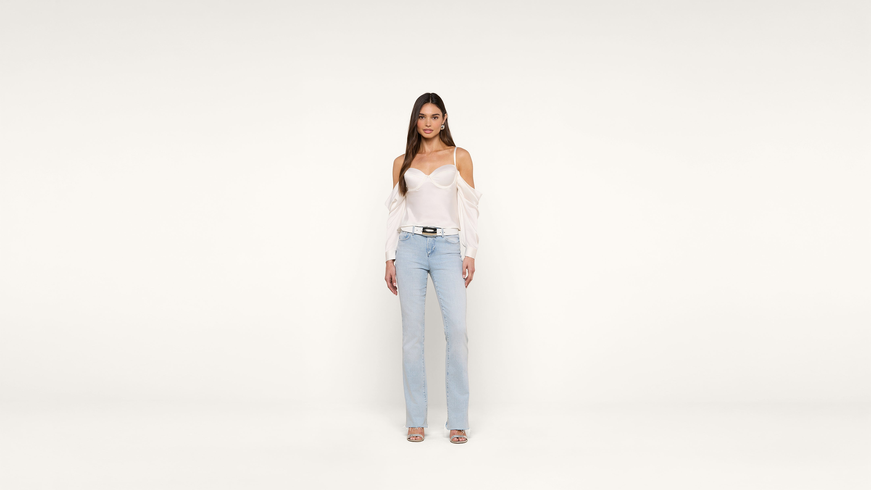 A woman stands against a plain white background, wearing a white off-shoulder blouse, light blue jeans, and heeled sandals. She looks directly at the camera with a neutral expression.