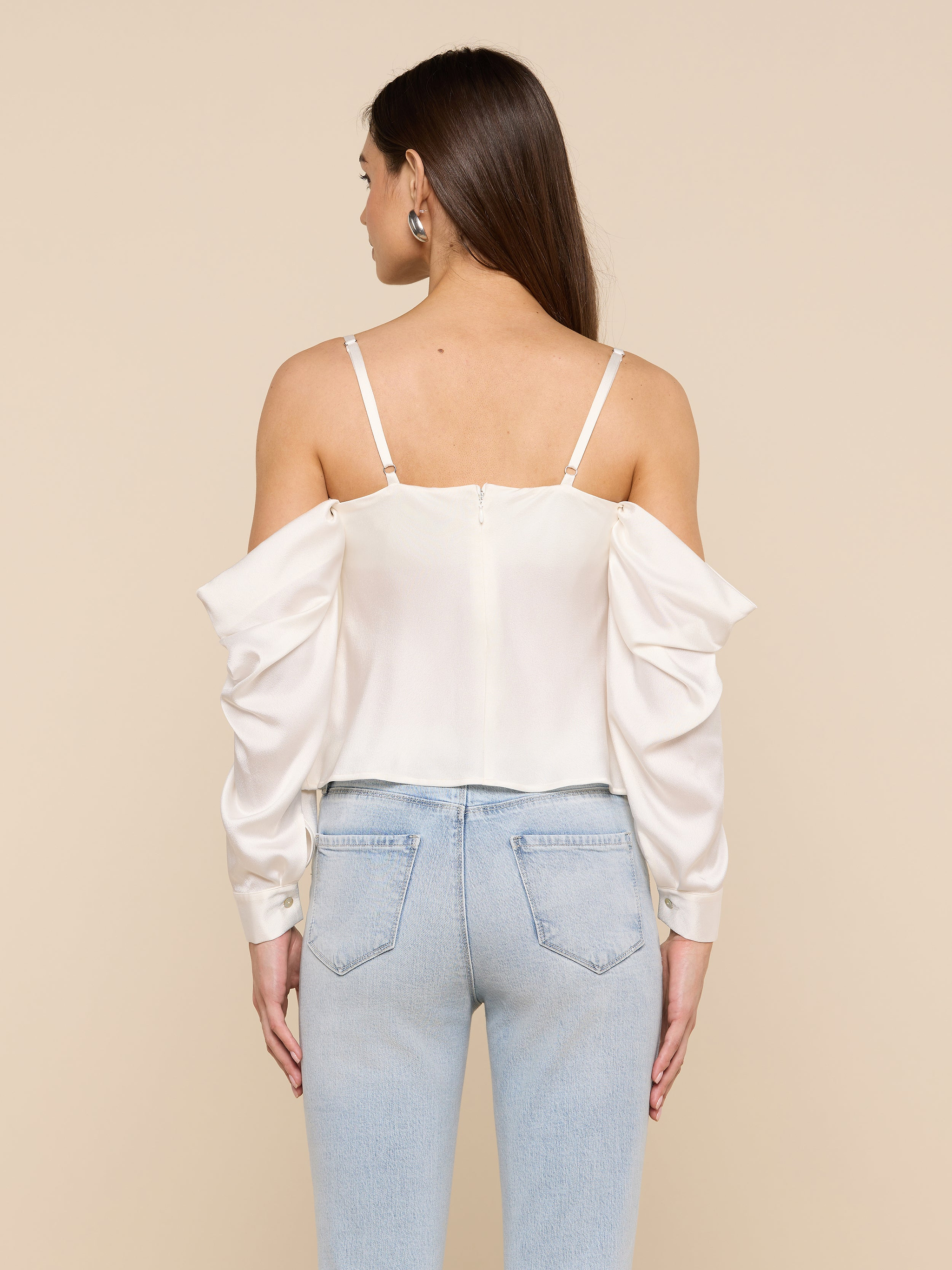 A woman is seen from behind wearing the Saya Satin Top—a white off-the-shoulder blouse with thin straps and long sleeves, styled with light blue high-waisted jeans against a plain light background. The Saya Satin Top offers a flattering fit for an elegant look.