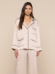 A woman stands facing forward, wearing the Mara Blouse — a cream-colored satin top with black piping and a relaxed fit button-up design. She pairs it with matching wide-leg pants, and stands against a plain light background.