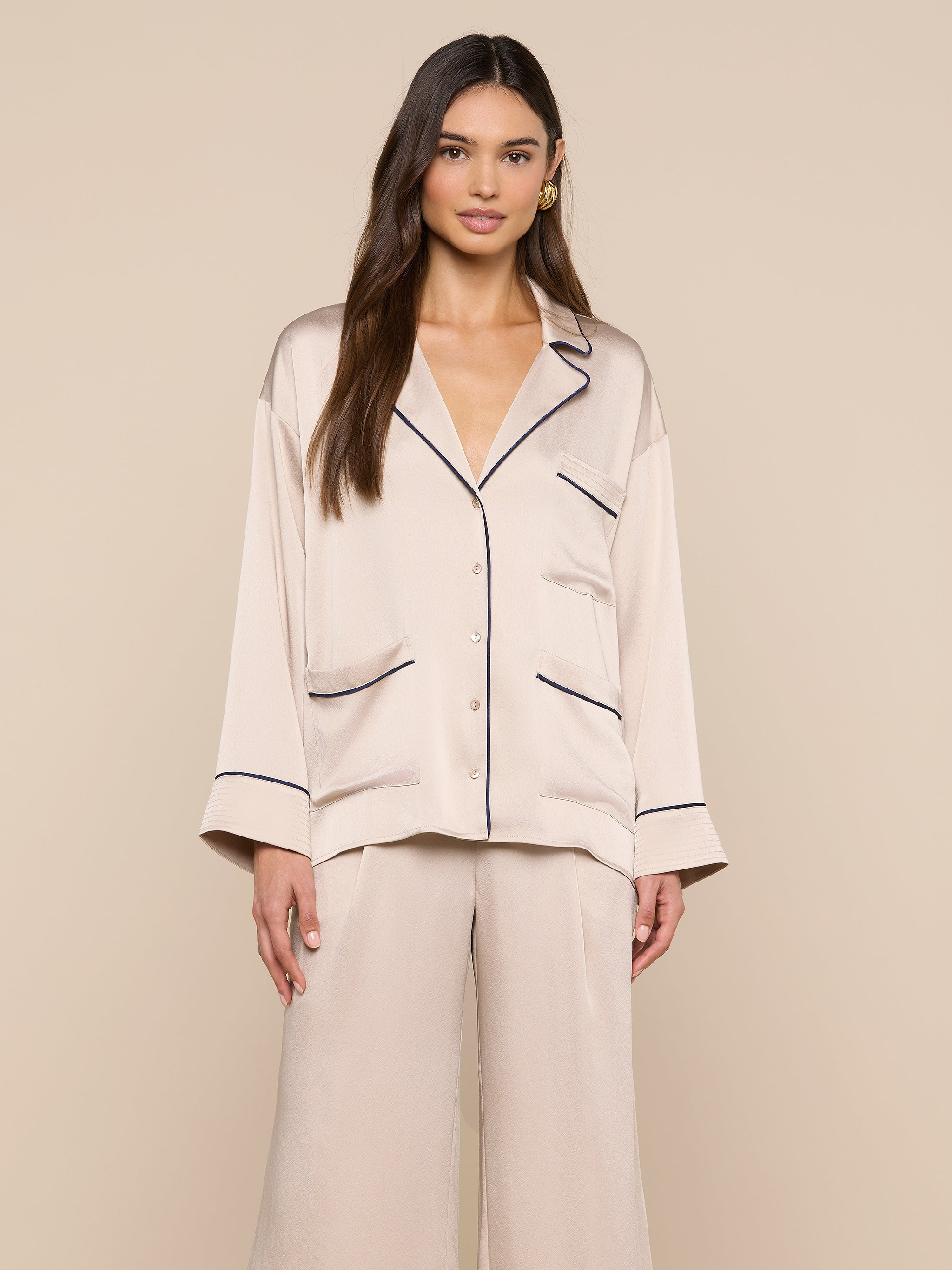 A woman stands facing forward, wearing the Mara Blouse — a cream-colored satin top with black piping and a relaxed fit button-up design. She pairs it with matching wide-leg pants, and stands against a plain light background.