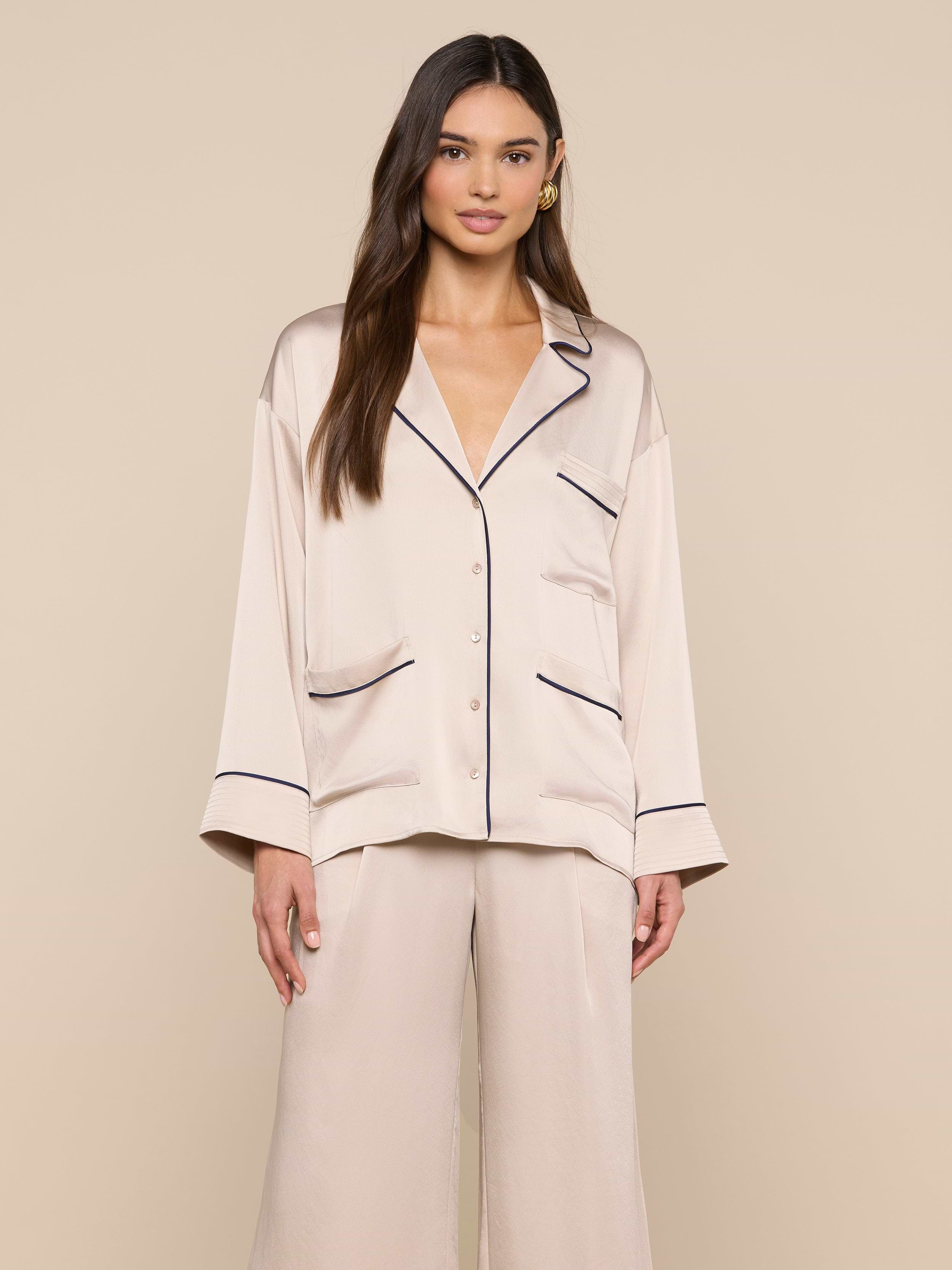 A woman stands facing forward, wearing the Mara Blouse — a cream-colored satin top with black piping and a relaxed fit button-up design. She pairs it with matching wide-leg pants, and stands against a plain light background.