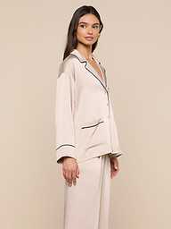 Model stands sideways, gazing slightly to the left, wearing the Mara Blouse in cream-colored satin with black trim and a relaxed fit. The plain, light background accentuates this elegant loungewear piece.
