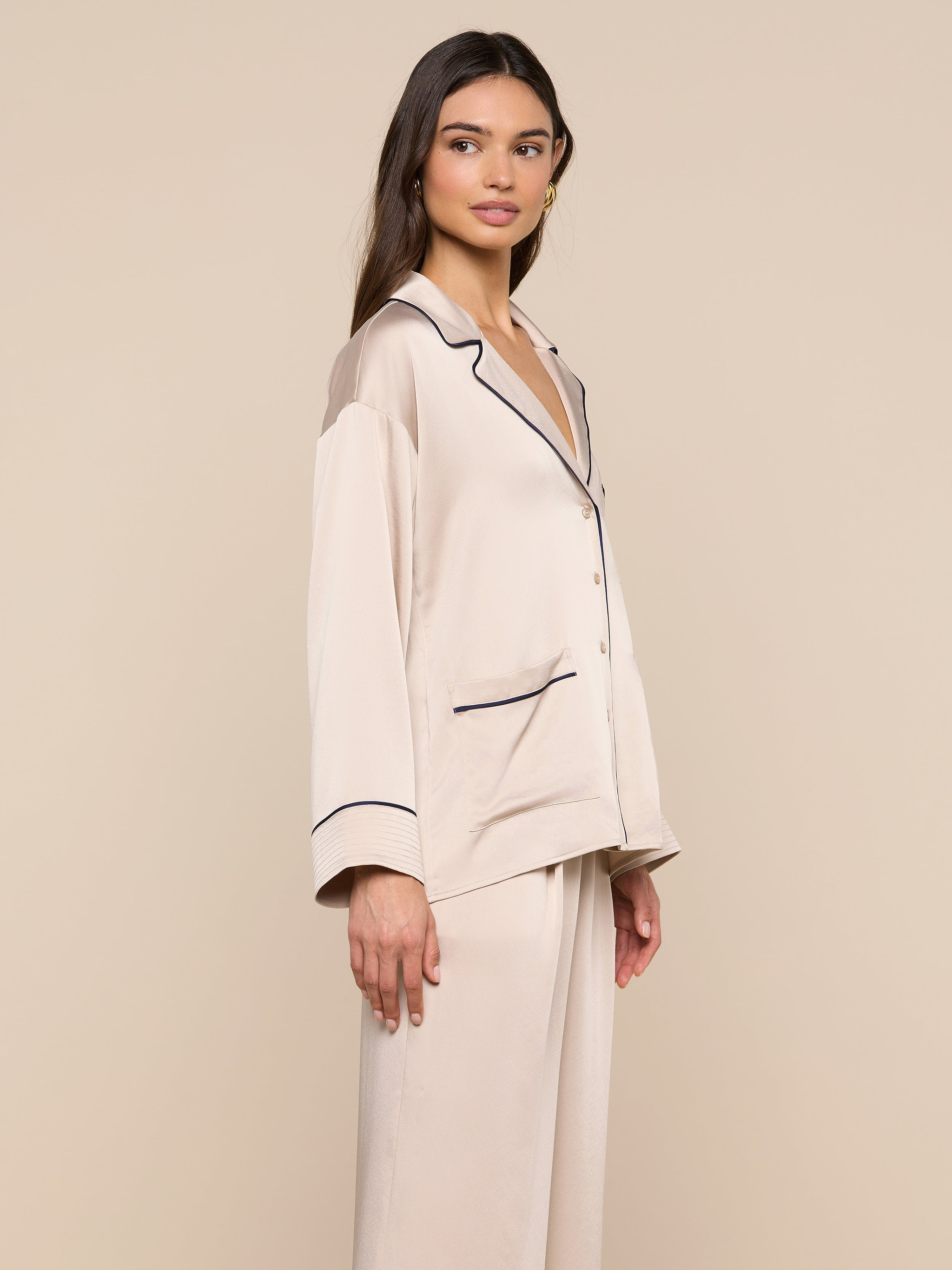 Model stands sideways, gazing slightly to the left, wearing the Mara Blouse in cream-colored satin with black trim and a relaxed fit. The plain, light background accentuates this elegant loungewear piece.