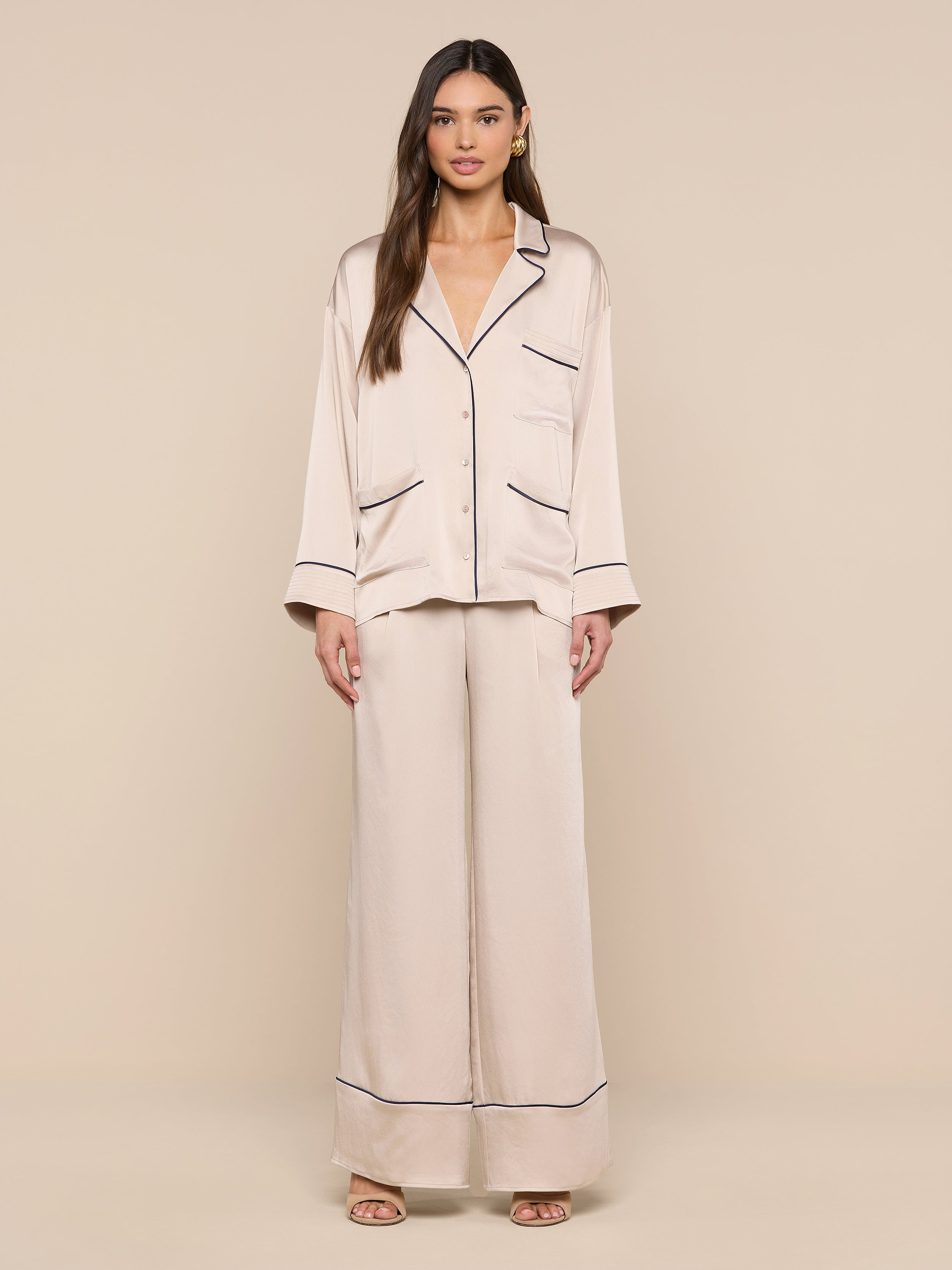 A woman stands facing forward, wearing the Mara Blouse in a relaxed fit light beige design with black piping details on the cuffs, pockets, and collar, against a plain white background.