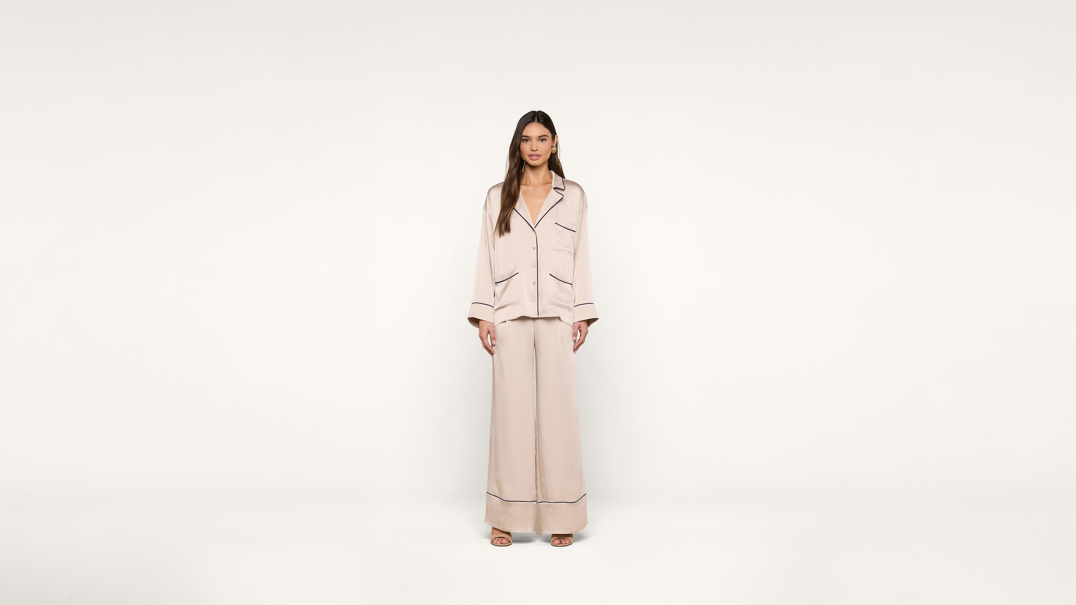 A woman stands against a plain white background, wearing a light beige pajama-style outfit with dark piping and open-toe shoes.