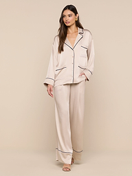 A woman stands against a plain white background, dressed in the Mara Blouse—a light beige satin top with dark trim—paired with open-toe sandals. The relaxed fit of the blouse enhances an effortless, comfortable style.