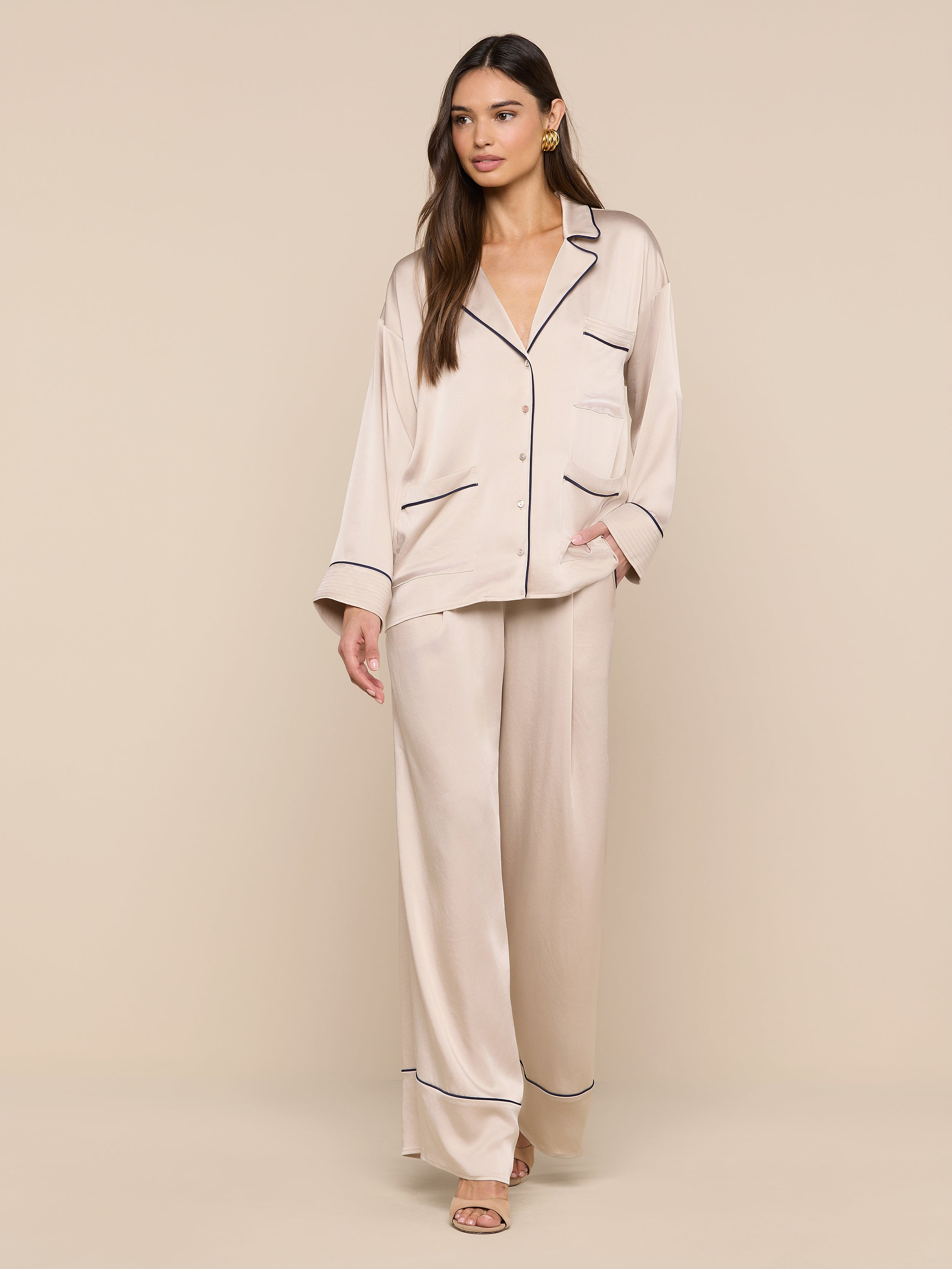A woman stands against a plain white background, dressed in the Mara Blouse—a light beige satin top with dark trim—paired with open-toe sandals. The relaxed fit of the blouse enhances an effortless, comfortable style.