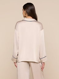 A woman with long brown hair is seen from behind, wearing the Mara Blouse—a relaxed fit, beige loungewear top with long sleeves and black piping details—standing against a plain light background.
