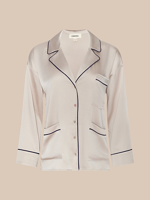 The Mara Blouse is a light pink satin long-sleeve pajama top with black piping, notched collar, relaxed fit, three front pockets, and button-down closure—ideal loungewear shown on a beige background.