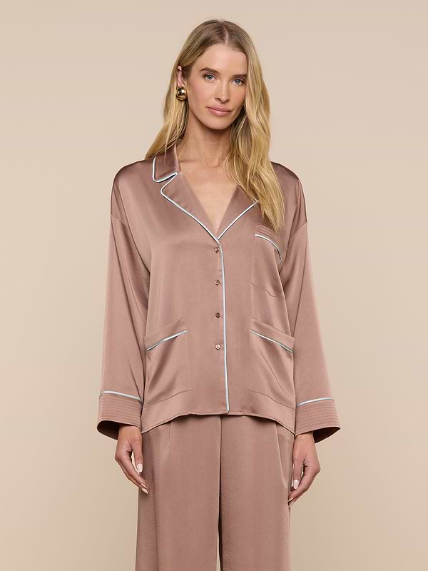 A woman wearing the Mara Blouse—a relaxed fit, silky, light brown top with white piping—stands against a plain light background, looking at the camera with a gentle smile.