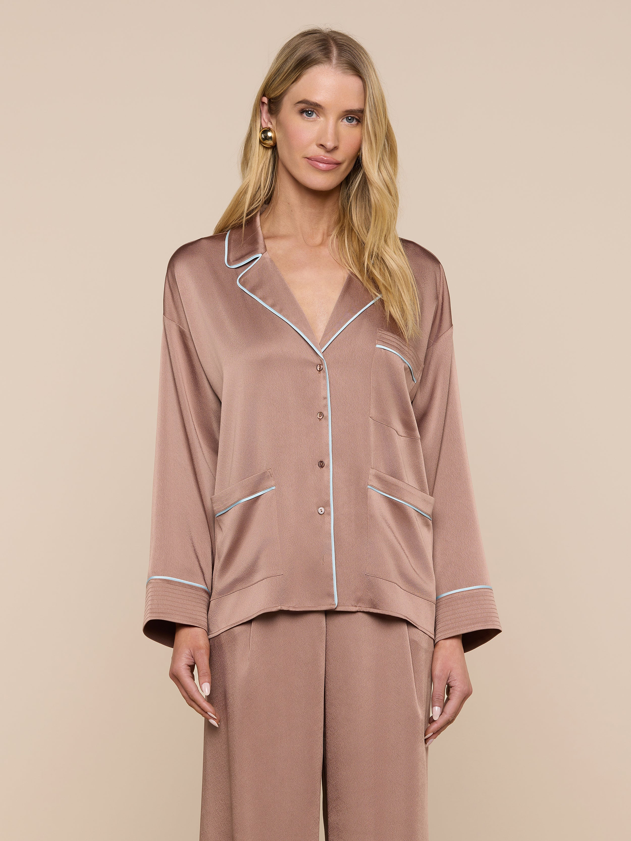 A woman wearing the Mara Blouse—a relaxed fit, silky, light brown top with white piping—stands against a plain light background, looking at the camera with a gentle smile.