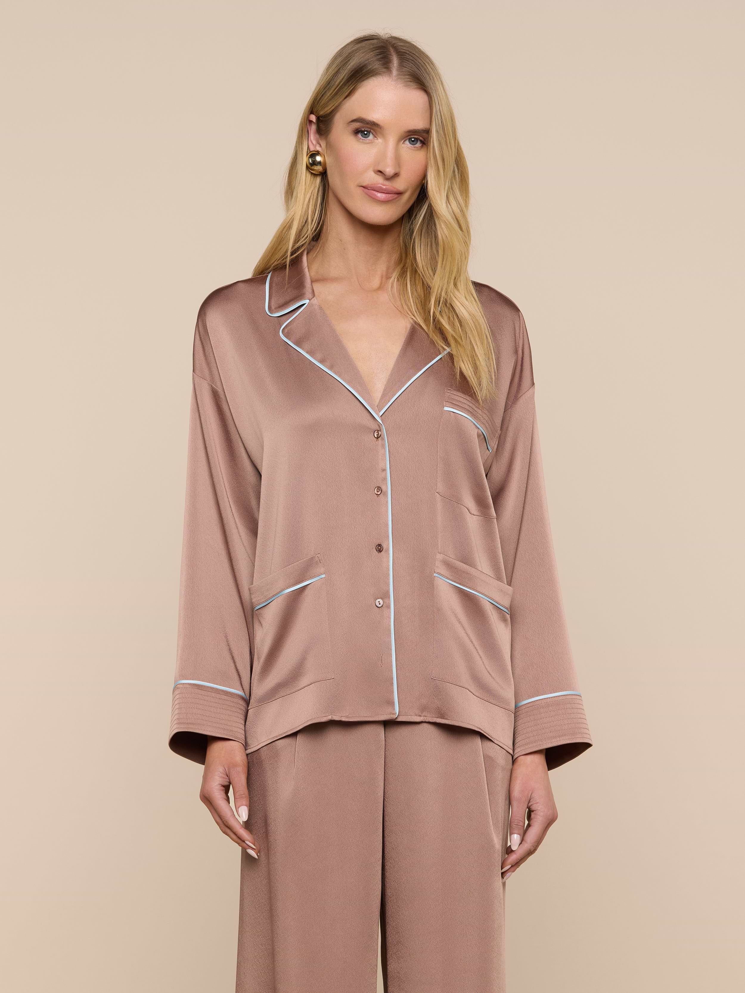 A woman wearing the Mara Blouse—a relaxed fit, silky, light brown top with white piping—stands against a plain light background, looking at the camera with a gentle smile.