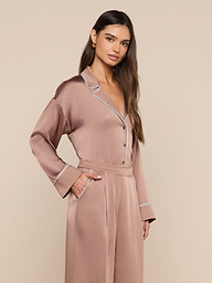 A woman stands against a plain background, wearing the Mara Blouse—a taupe-colored top with white piping. She has her hands in the blouse’s pockets and her long brown hair is styled loosely over one shoulder, while the relaxed fit adds to its effortless appeal.