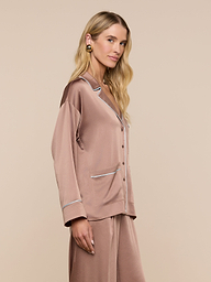 A woman with long blonde hair wears the Mara Blouse, a mauve-colored loungewear top featuring white piping and a relaxed fit. She stands sideways, looking forward against a plain, light background.