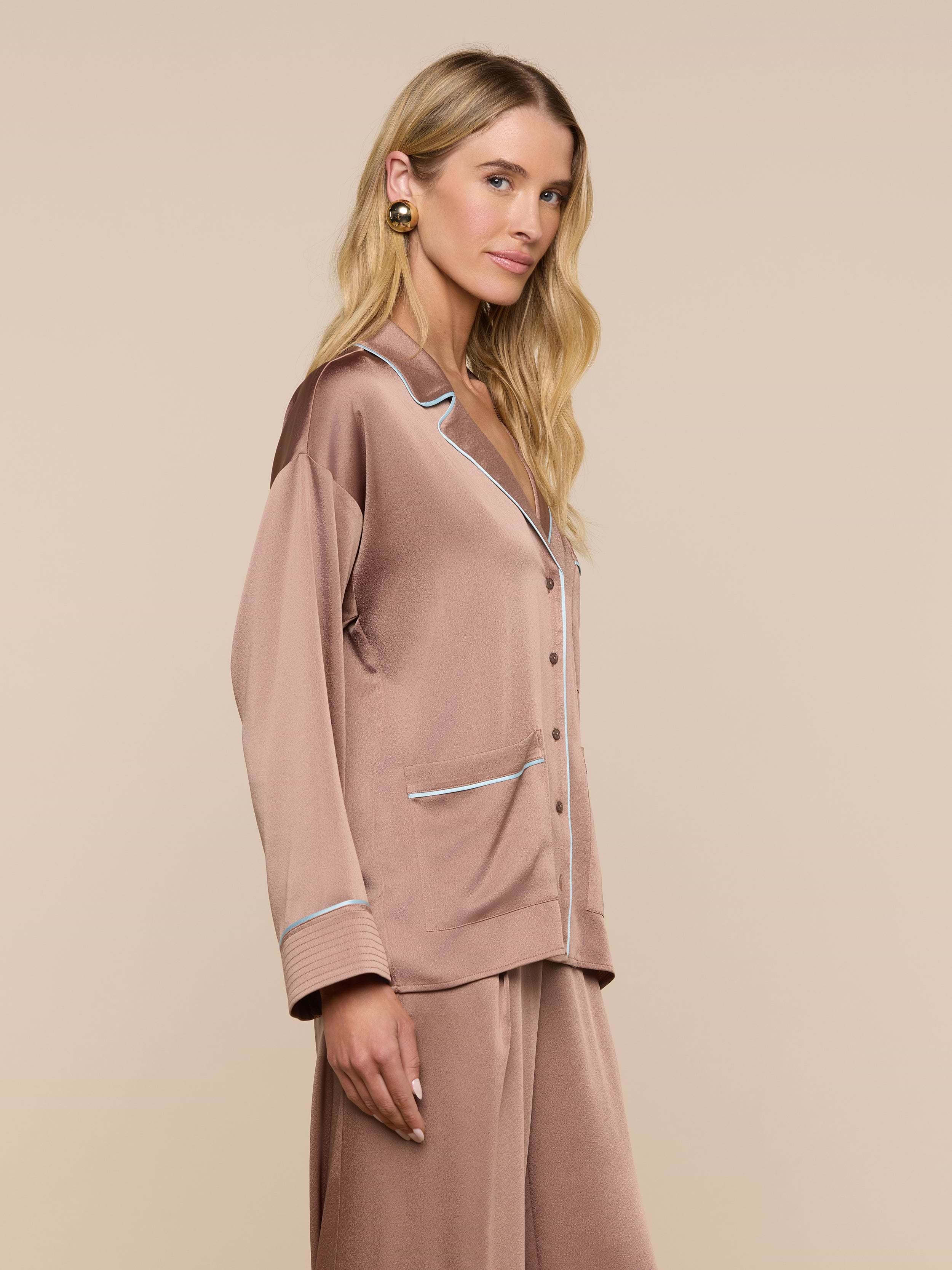 A woman with long blonde hair wears the Mara Blouse, a mauve-colored loungewear top featuring white piping and a relaxed fit. She stands sideways, looking forward against a plain, light background.