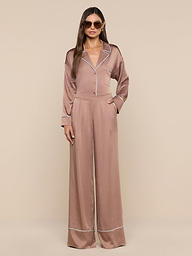 A woman wearing sunglasses and the Mara Blouse in light brown satin with white piping stands against a plain white background, showcasing confident loungewear in a relaxed fit, one hand in her pocket.