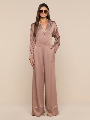 A woman in sunglasses and the Mara Blouse—with a silky taupe finish, white piping, long sleeves, and relaxed fit—poses against a beige background, channeling effortless loungewear with her hands in her pockets.
