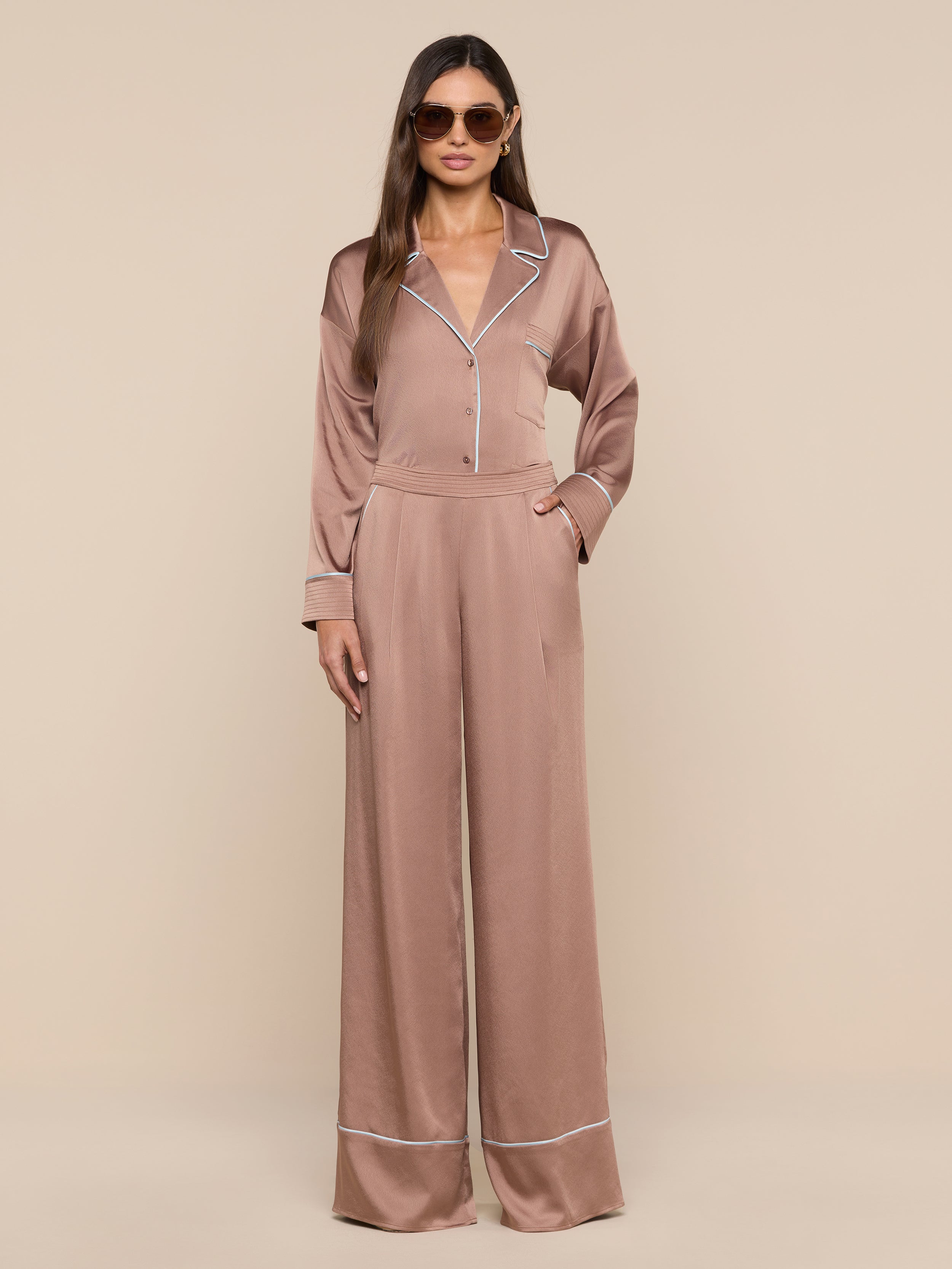 A woman wearing sunglasses and the Mara Blouse in light brown satin with white piping stands against a plain white background, showcasing confident loungewear in a relaxed fit, one hand in her pocket.