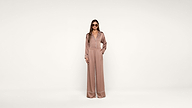 A woman wearing sunglasses and a pink, long-sleeved, wide-leg jumpsuit with white piping stands against a plain white background, hands in pockets.
