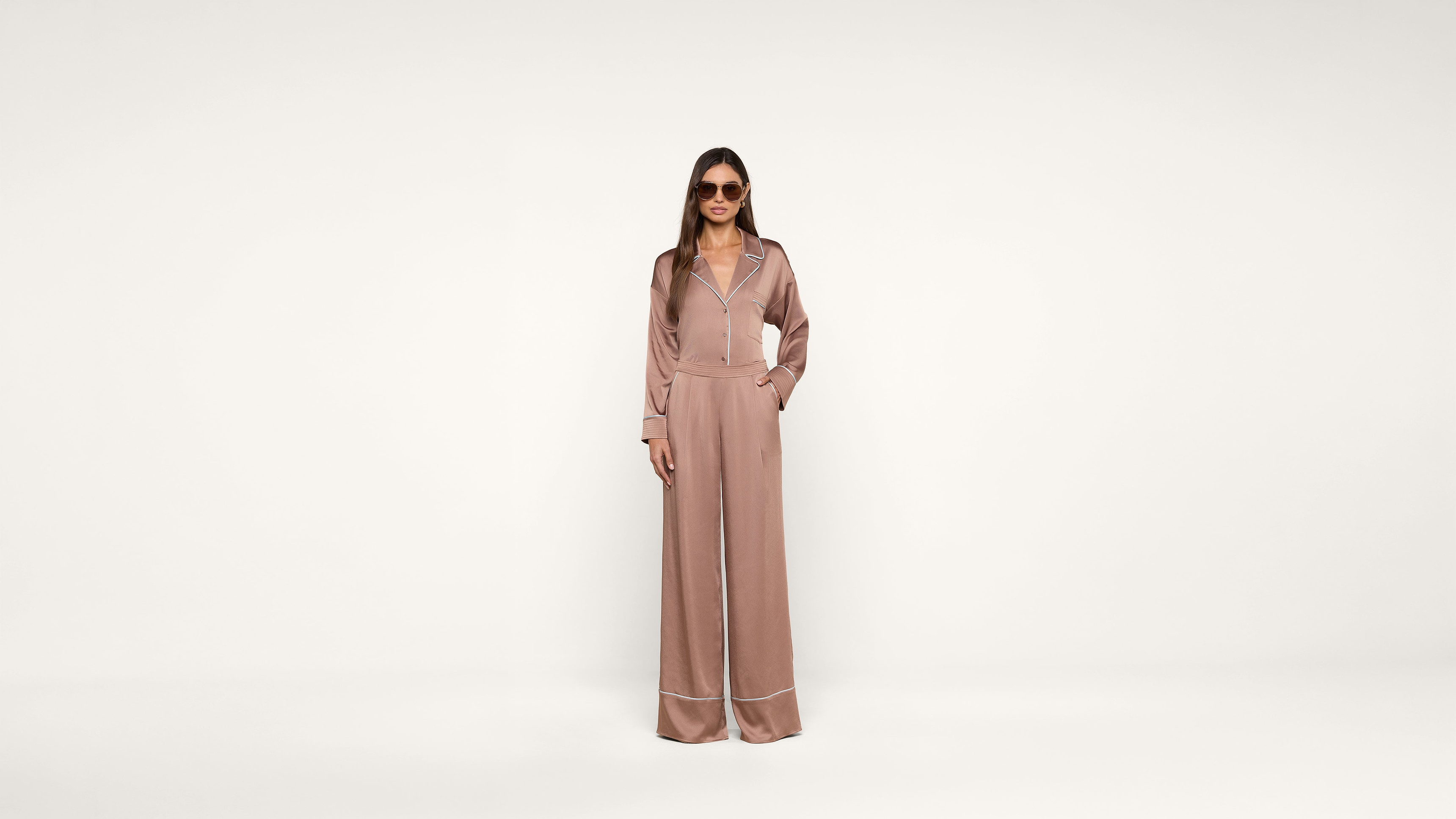 A woman wearing sunglasses and a pink, long-sleeved, wide-leg jumpsuit with white piping stands against a plain white background, hands in pockets.