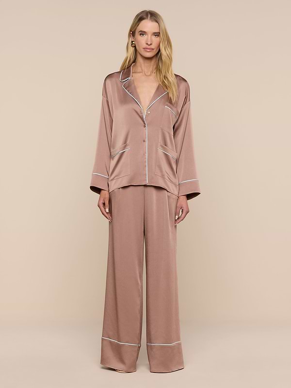 A woman with long blonde hair and a neutral expression stands against a plain background wearing the Mara Blouse—a relaxed-fit, silky brown button-up top with white trim—paired with matching wide-leg pajama pants.