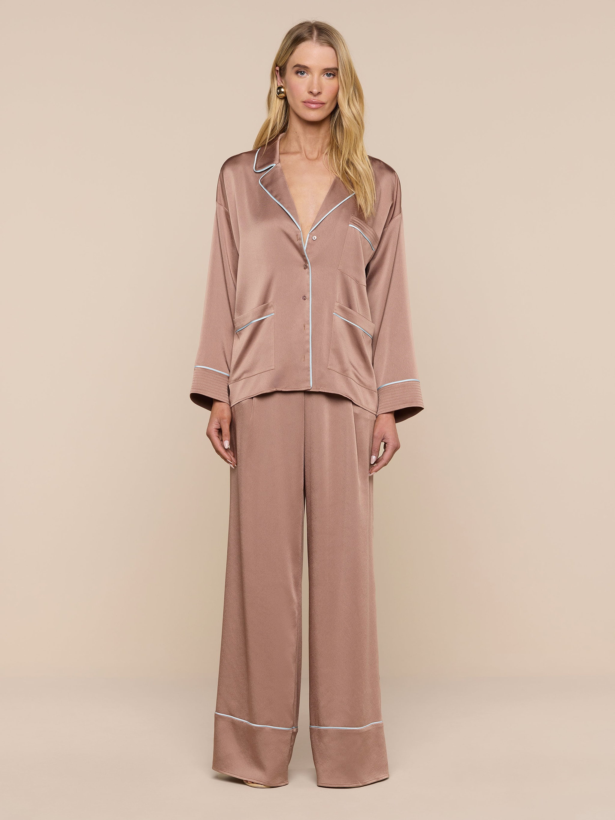 A woman with long blonde hair and a neutral expression stands against a plain background wearing the Mara Blouse—a relaxed-fit, silky brown button-up top with white trim—paired with matching wide-leg pajama pants.