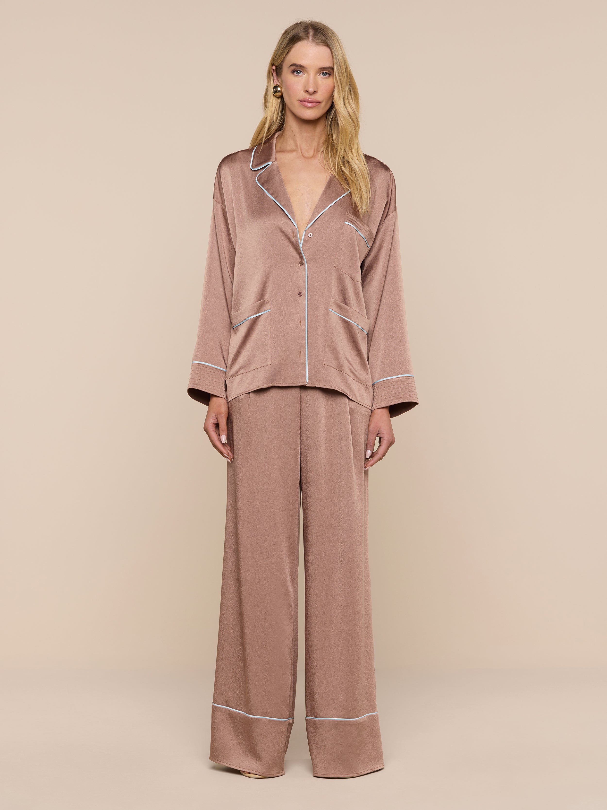 A woman with long blonde hair and a neutral expression stands against a plain background wearing the Mara Blouse—a relaxed-fit, silky brown button-up top with white trim—paired with matching wide-leg pajama pants.