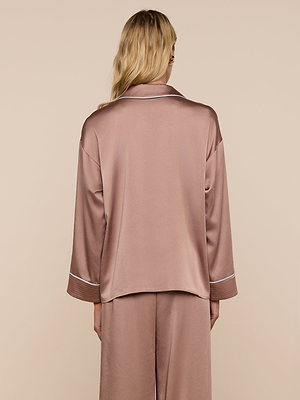 A person with long blonde hair is shown from the back, wearing the Mara Blouse—a relaxed fit, silky, rose-colored top with white piping on the cuffs and collar—set against a plain beige background.