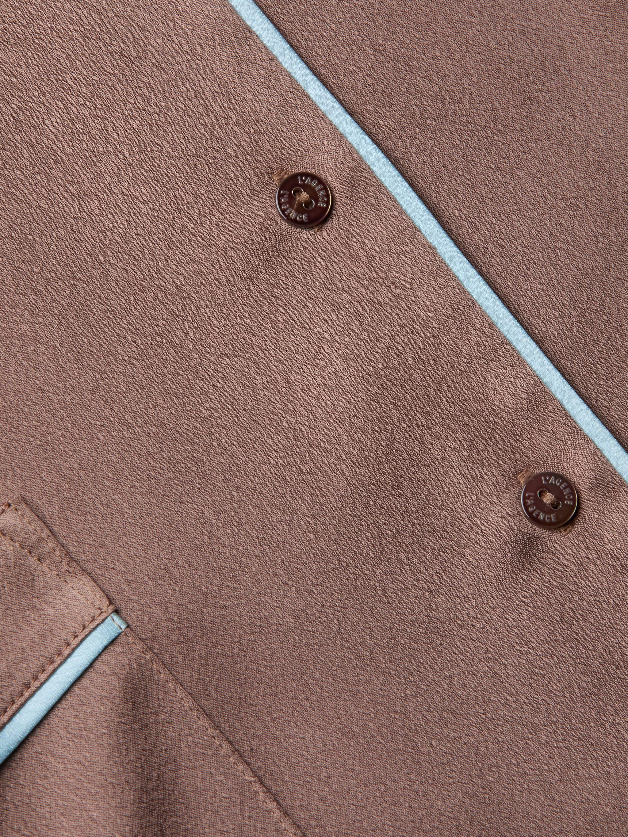 Close-up of the Mara Blouse in brown fabric, featuring two buttons embossed with LAINE ET CACHE, light blue piping along the edges, and a corner pocket accented with matching blue trim. The relaxed fit and thoughtful detailing evoke the refined comfort of premium loungewear.