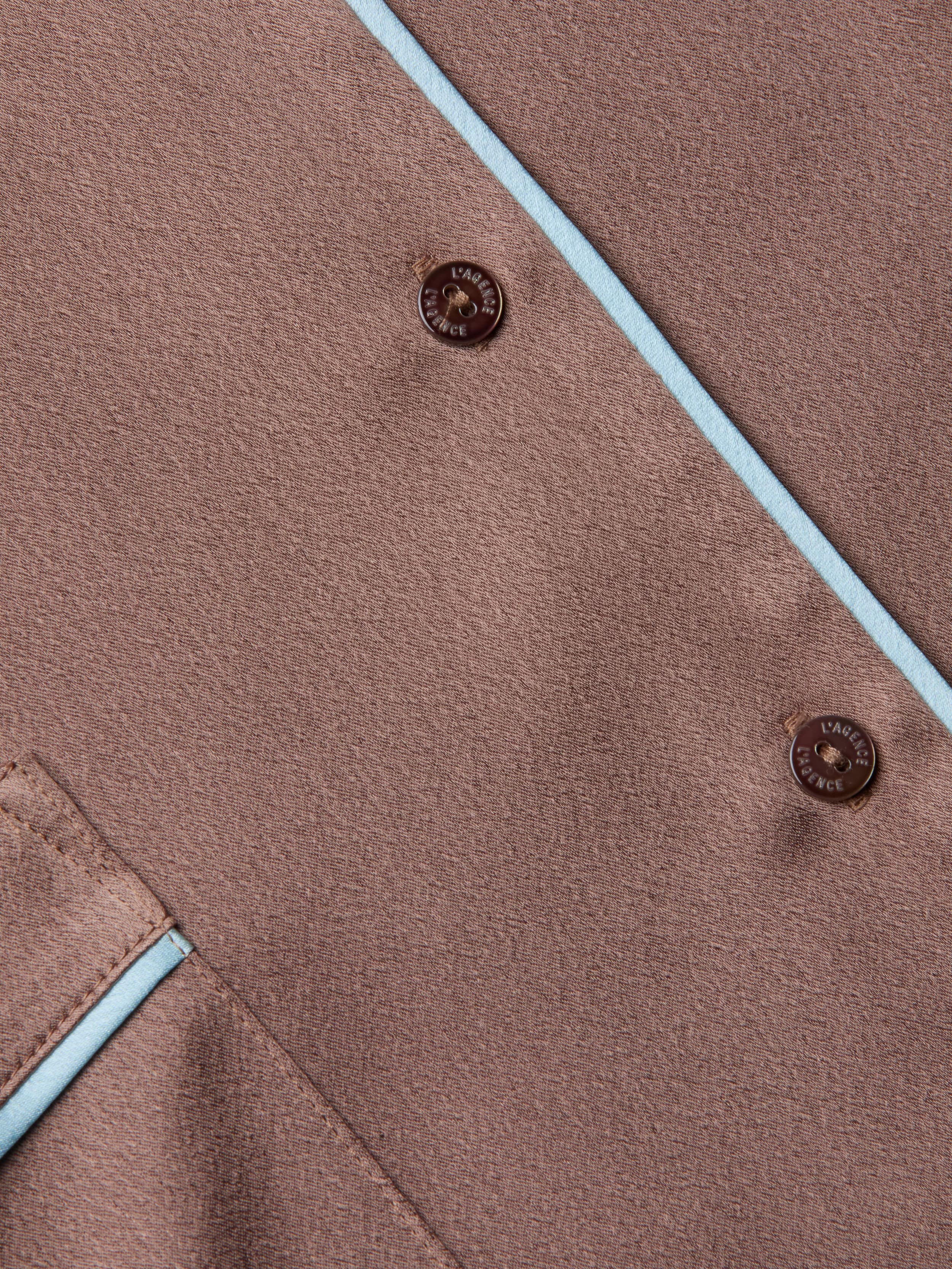 Close-up of the Mara Blouse in brown fabric, featuring two buttons embossed with LAINE ET CACHE, light blue piping along the edges, and a corner pocket accented with matching blue trim. The relaxed fit and thoughtful detailing evoke the refined comfort of premium loungewear.