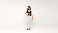 A woman wearing sunglasses, a black lace top, white pants, and black heels stands confidently against a plain white background.