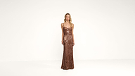 A woman stands against a plain white background, wearing a long, bronze-colored, sequin evening gown with thin straps.
