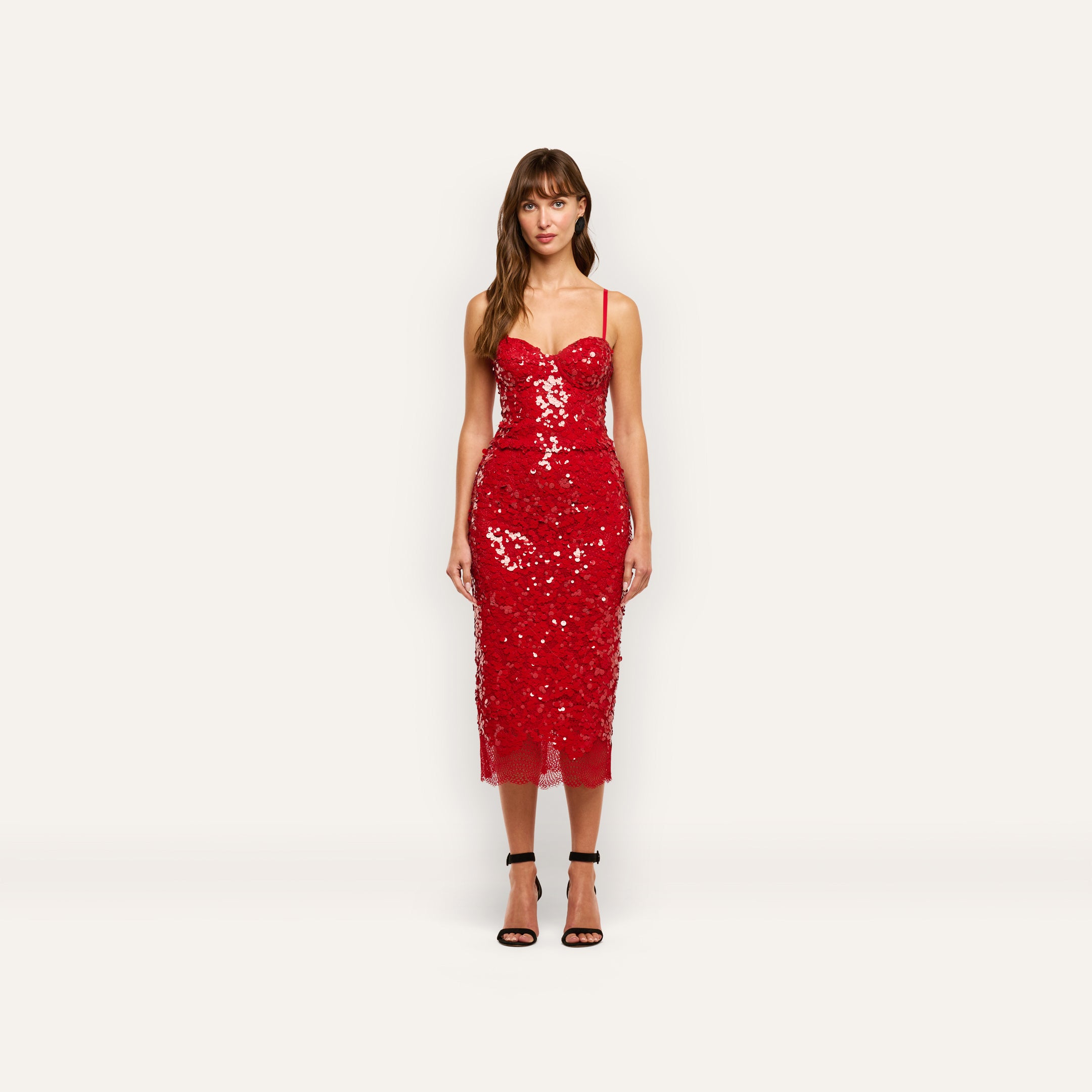 A model wearing the Lior Sequin Top as a red, spaghetti-strap sequined midi dress with a structured, bustier-style bodice and a sheer scalloped hem.