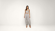 A woman stands in a minimal studio wearing a light grey sleeveless crop top and loose grey pants with gathered ankles, paired with strappy heels.