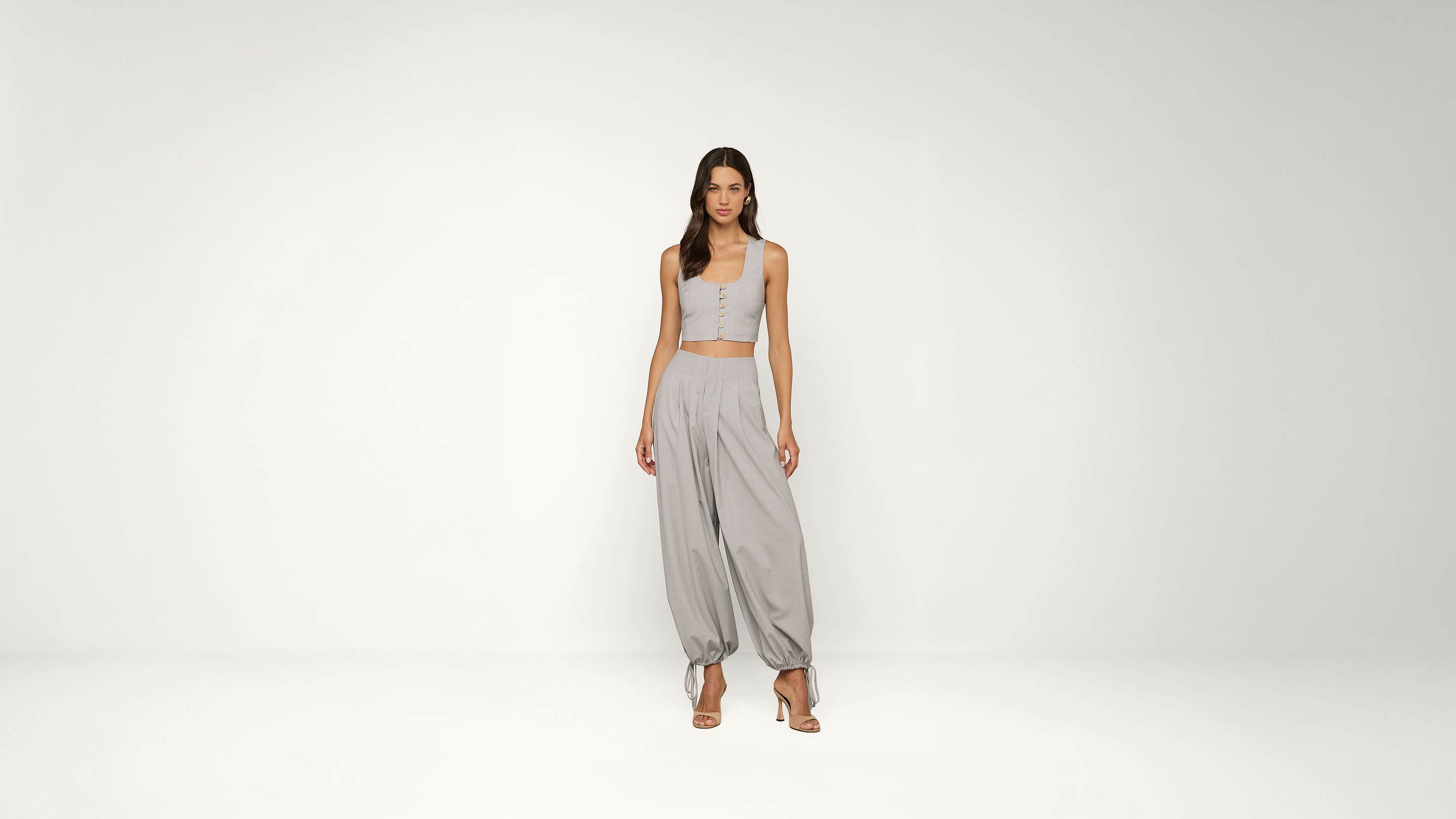 A woman stands in a minimal studio wearing a light grey sleeveless crop top and loose grey pants with gathered ankles, paired with strappy heels.