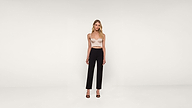 A woman stands against a plain white background wearing a light-colored cropped top, high-waisted black pants, and black high-heeled sandals.