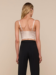 A woman with long blonde hair, seen from behind, wears the Nysa Bustier Top—a beige sleeveless crop top with adjustable straps—paired with high-waisted black pants against a plain background.