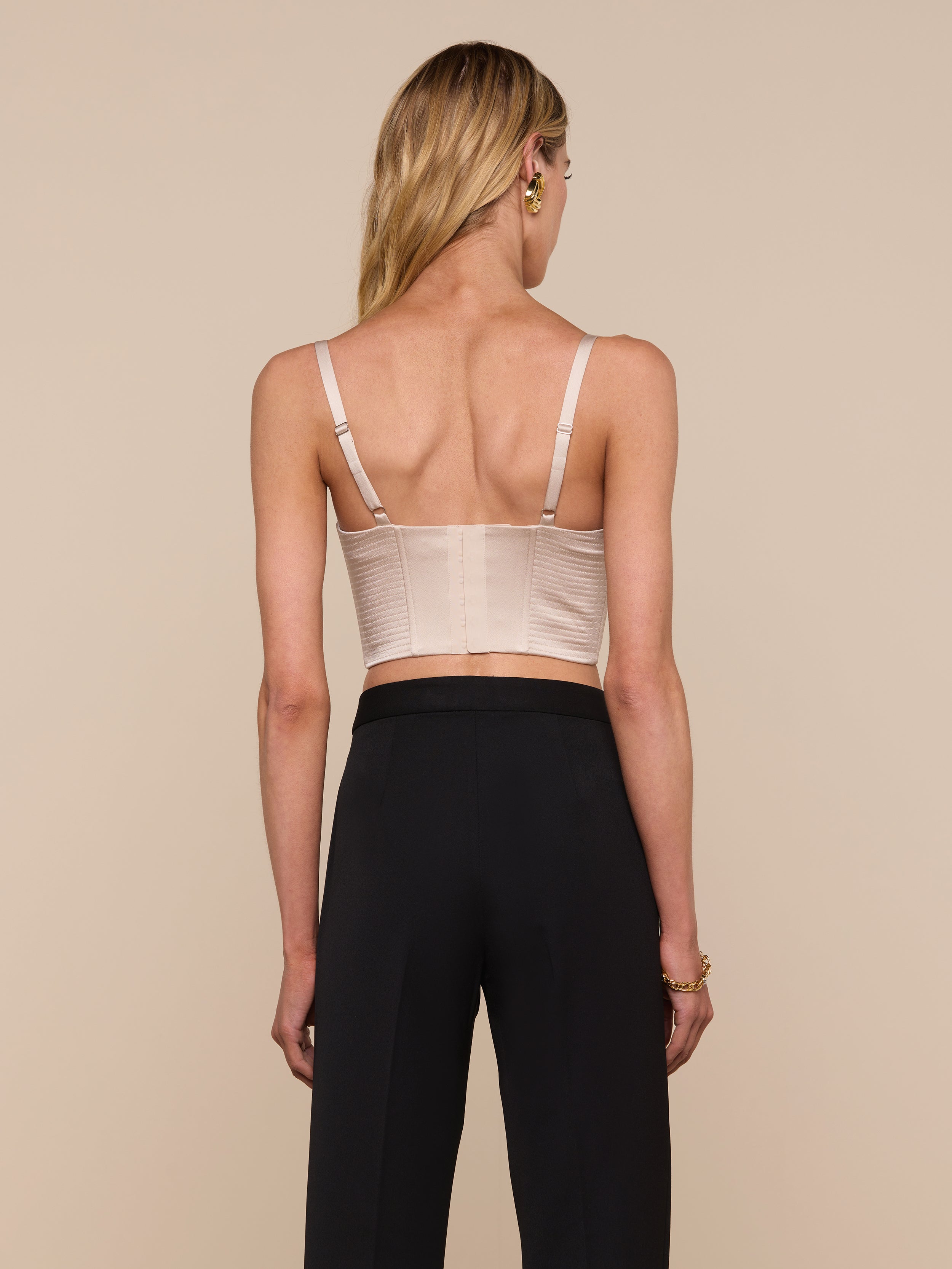 A woman with long blonde hair, seen from behind, wears the Nysa Bustier Top—a beige sleeveless crop top with adjustable straps—paired with high-waisted black pants against a plain background.