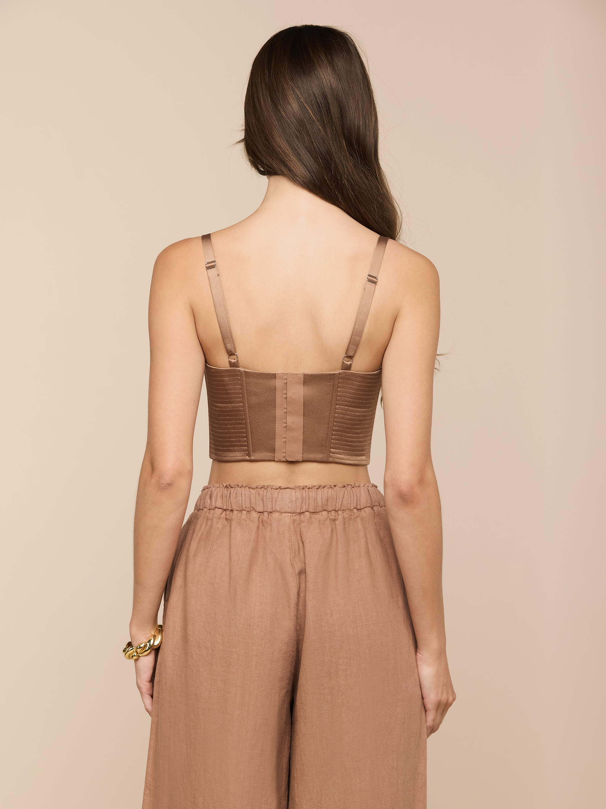 A woman with long brown hair faces away, wearing the Nysa Bustier Top—a brown cropped top with adjustable straps—and loose brown pants. She has a gold bracelet on her left wrist. The background is plain and light.