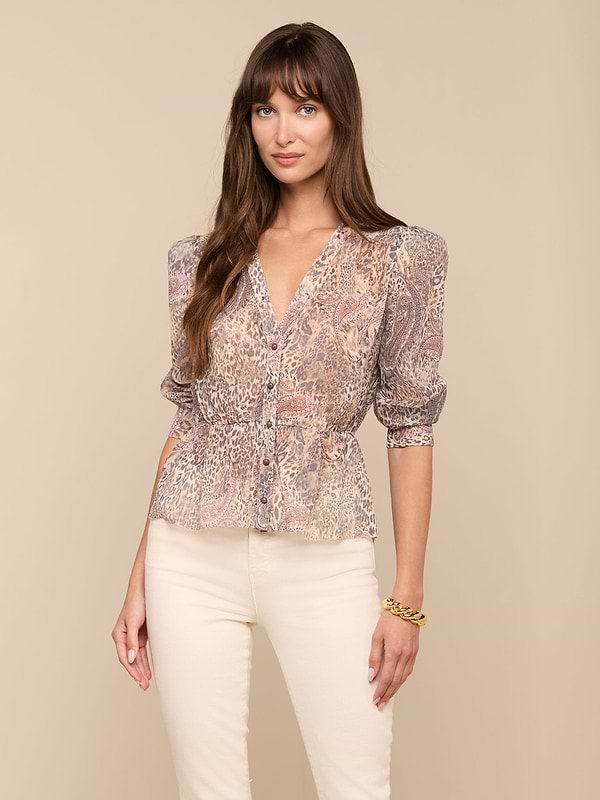 A woman with long brown hair wears the Suri Top with a paisley animal print and puff sleeves, paired with cream pants. She stands against a plain light background, looking at the camera with a slight smile.
