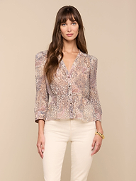 A woman with long brown hair wears the Suri Top—a sheer poly chiffon blouse featuring puff sleeves and a paisley animal print—paired with light-colored pants, standing against a plain light background.