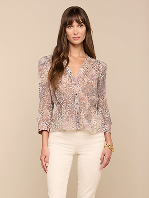 A woman with long brown hair wears the Suri Top, a patterned long-sleeve blouse, paired with cream-colored pants. She stands against a plain beige background, smiling softly with one hand by her side.