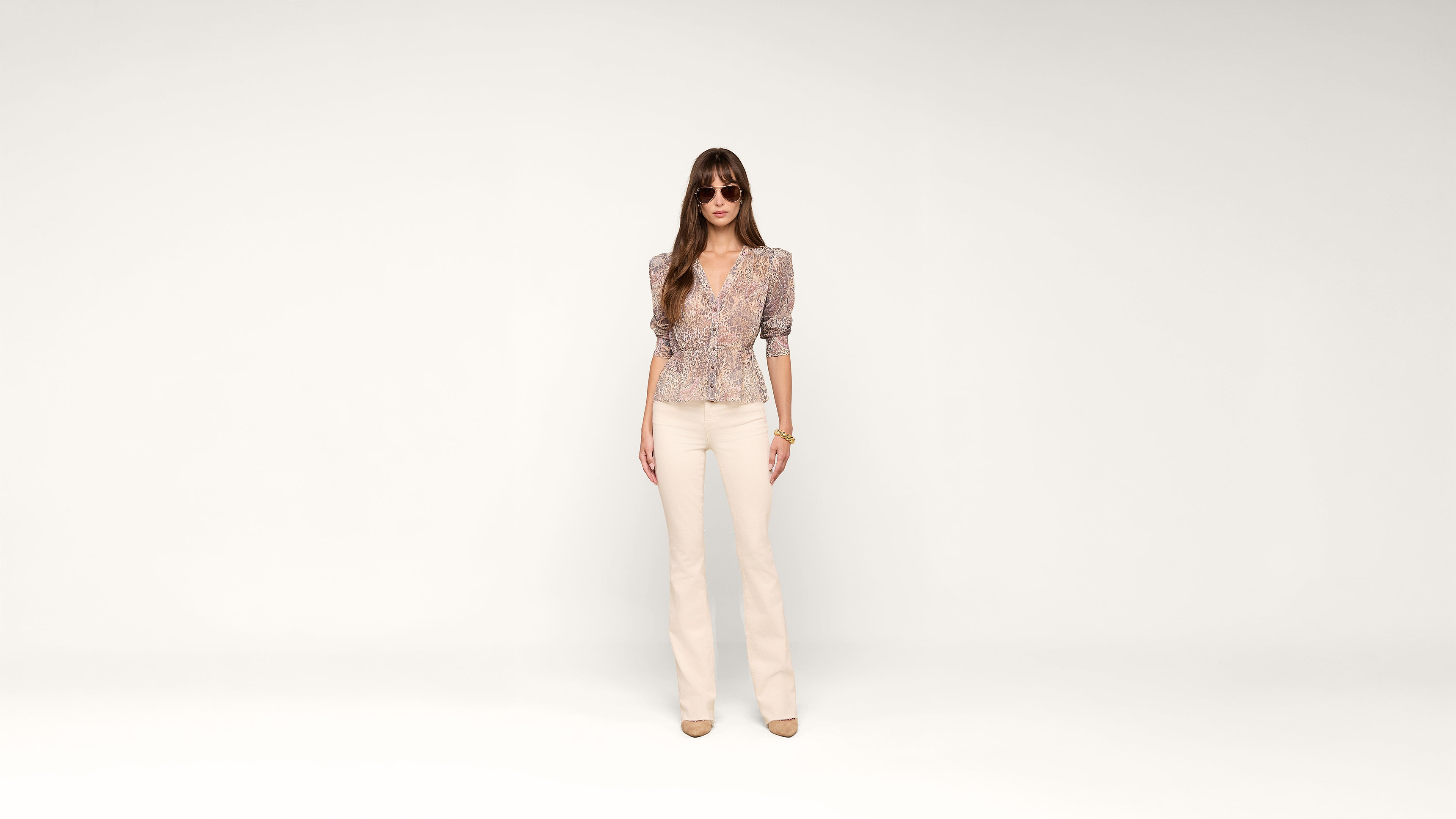 A woman wearing sunglasses, a patterned blouse, light-colored pants, and beige heels stands against a plain white background.