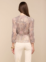 A woman with long brown hair, wearing the Suri Top—a sheer paisley animal print blouse with three-quarter sleeves—and cream-colored pants stands facing away from the camera against a plain background.
