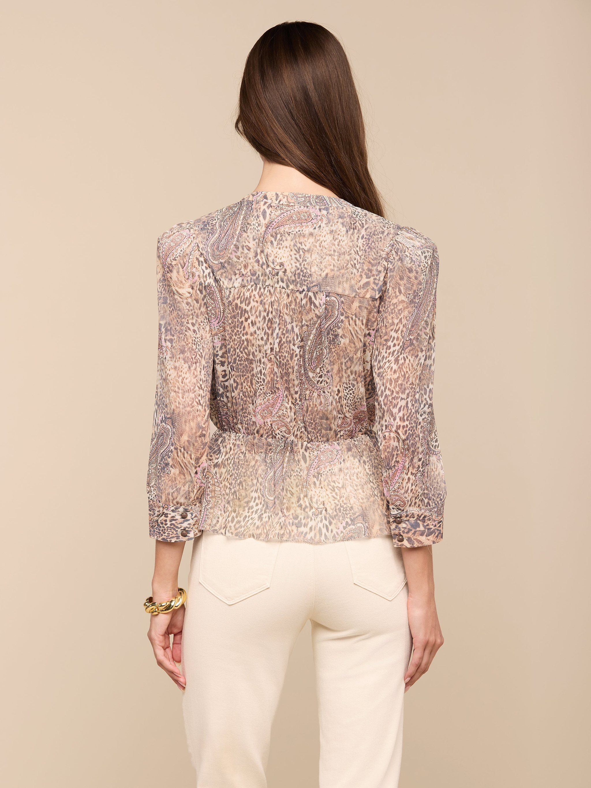 A woman with long brown hair, wearing the Suri Top—a sheer paisley animal print blouse with three-quarter sleeves—and cream-colored pants stands facing away from the camera against a plain background.
