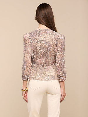 A woman with long brown hair, seen from behind against a beige background, wears the Suri Top—a sheer animal-print blouse with long sleeves—paired with light-colored high-waisted pants.
