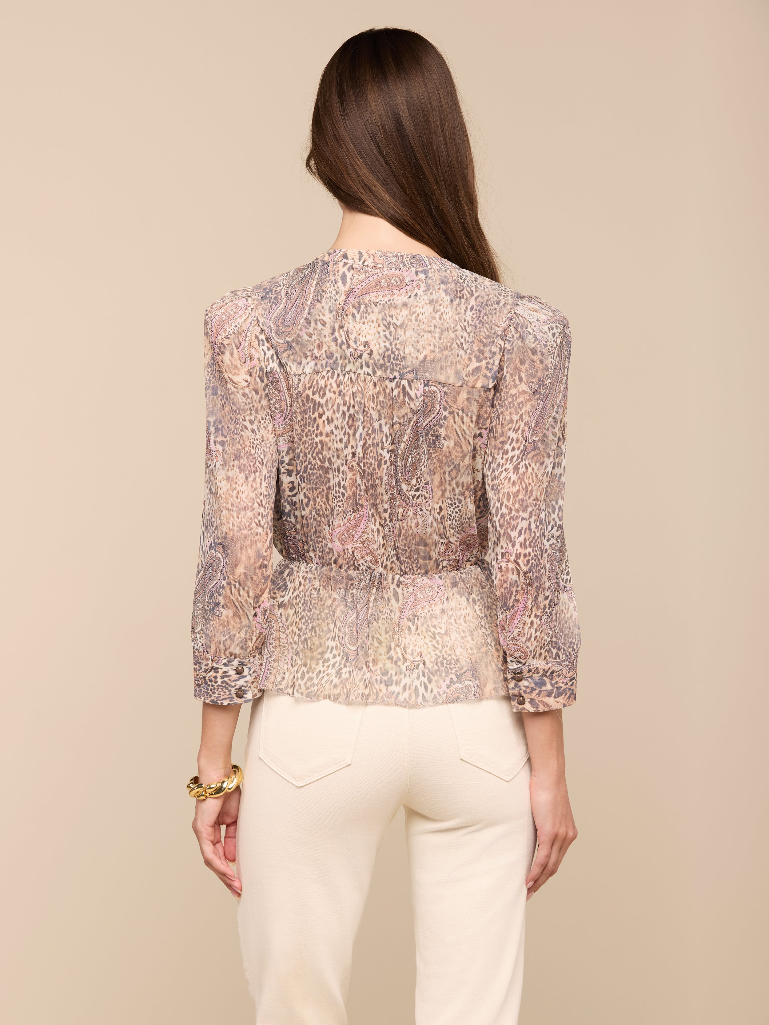 A woman with long brown hair, wearing the Suri Top—a sheer paisley animal print blouse with three-quarter sleeves—and cream-colored pants stands facing away from the camera against a plain background.
