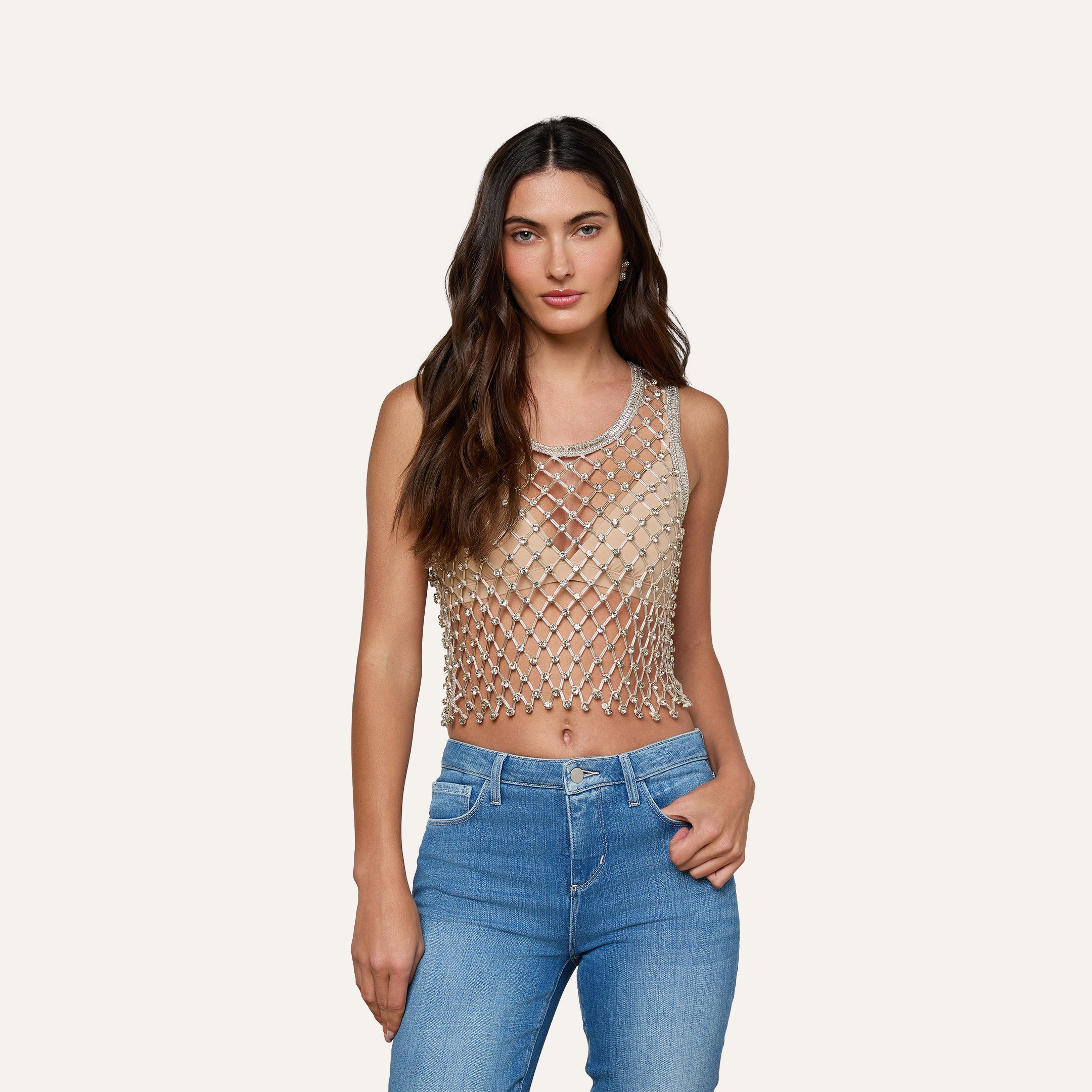 A woman with long brown hair wears the Freia Crystal Tank, featuring an artisanal beaded design with a diamond-patterned net overlay, paired with blue jeans and standing against a plain light background.